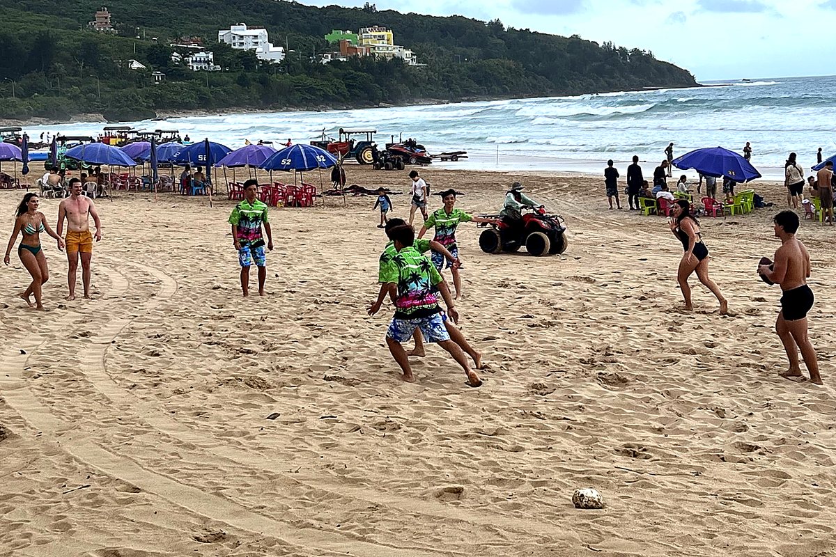 The group spread out across the sand mid-play, one person holding the football, waves breaking behind.