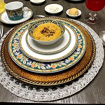 A bowl of golden squash soup topped with nuts, on a floral plate and gold charger.